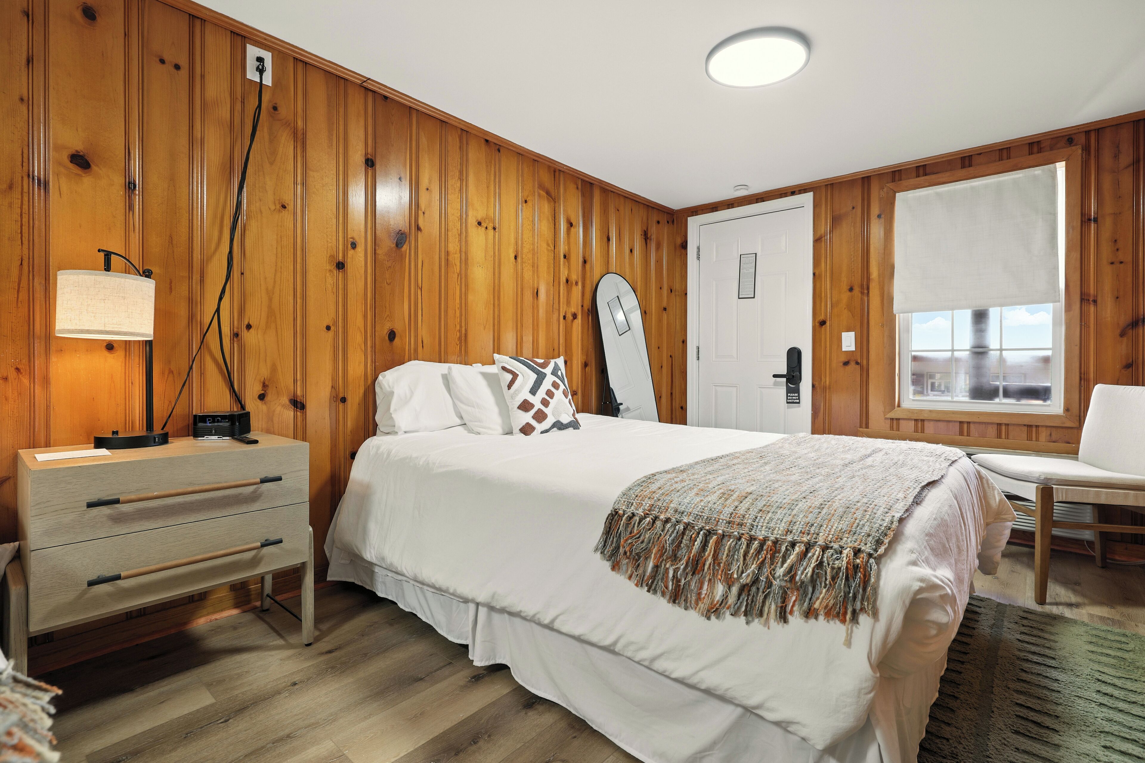 A bedroom with a wooden paneled wall, a bed with white bedding, a bedside table with a lamp, a chair, and a window with a view.