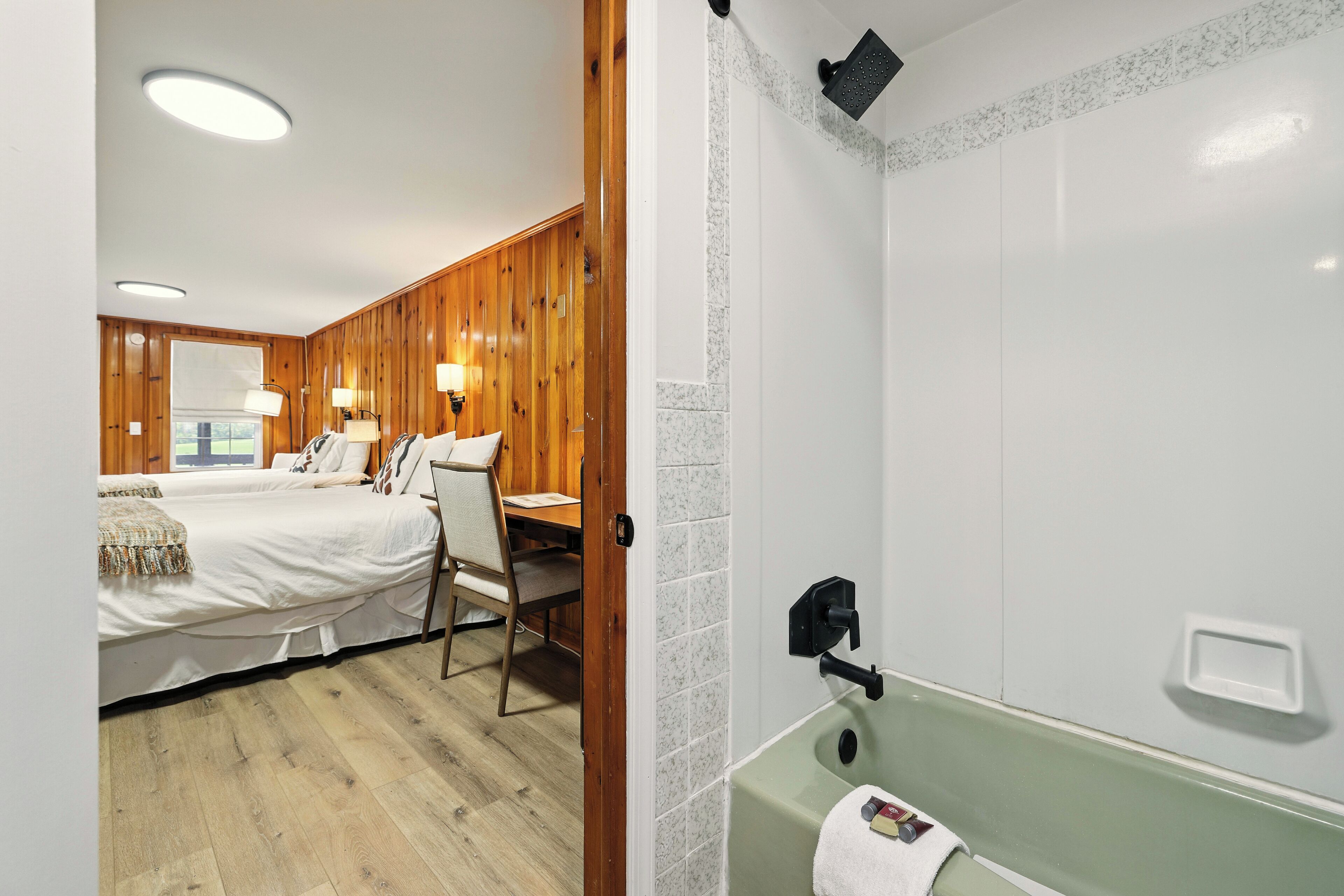 A room with two beds, a wooden wall, a green bathtub, and a window with a view.