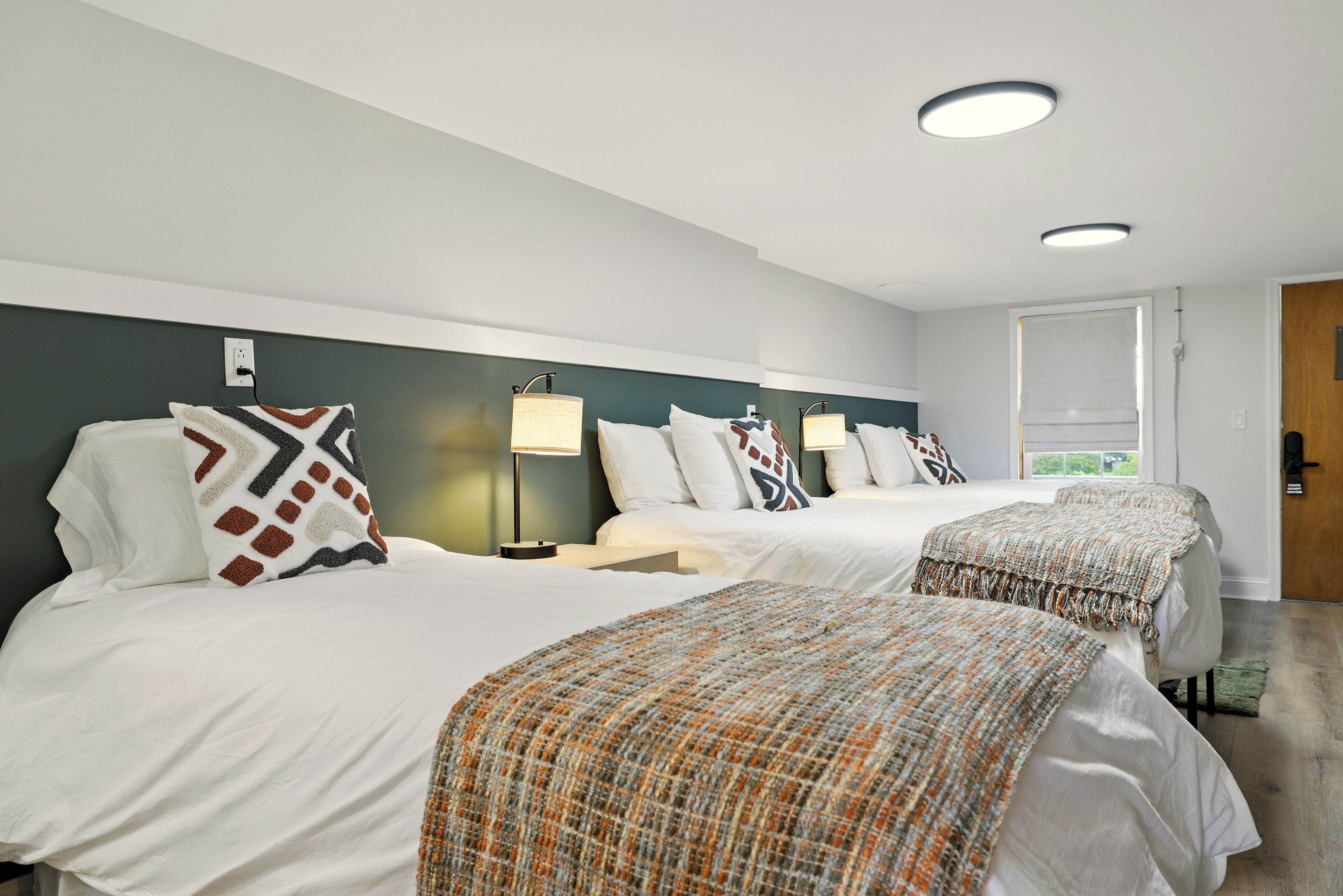 A hotel room with two beds, each with white bedding and patterned throw pillows. There are bedside lamps and a window with a view.