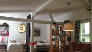 Amplia sala de estar con un sofá, una mesa de centro y una estantería. Se observa una cama con cabecera y una ventana con cortinas.