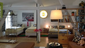 Una sala de estar acogedora con un sofá, una mesa de centro y una estantería. Se observa una cama con dosel y una lámpara sobre una mesita de noche.