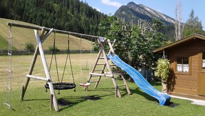 Ein Spielplatz mit einer Holzschaukel, einer Rutsche und einem kleinen Holzhaus, vor einer Berglandschaft.