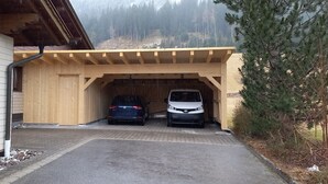 Ein überdachter Parkbereich mit zwei Autos, einer Holzhütte und einem Gebäude mit weißen Ziegelwänden.