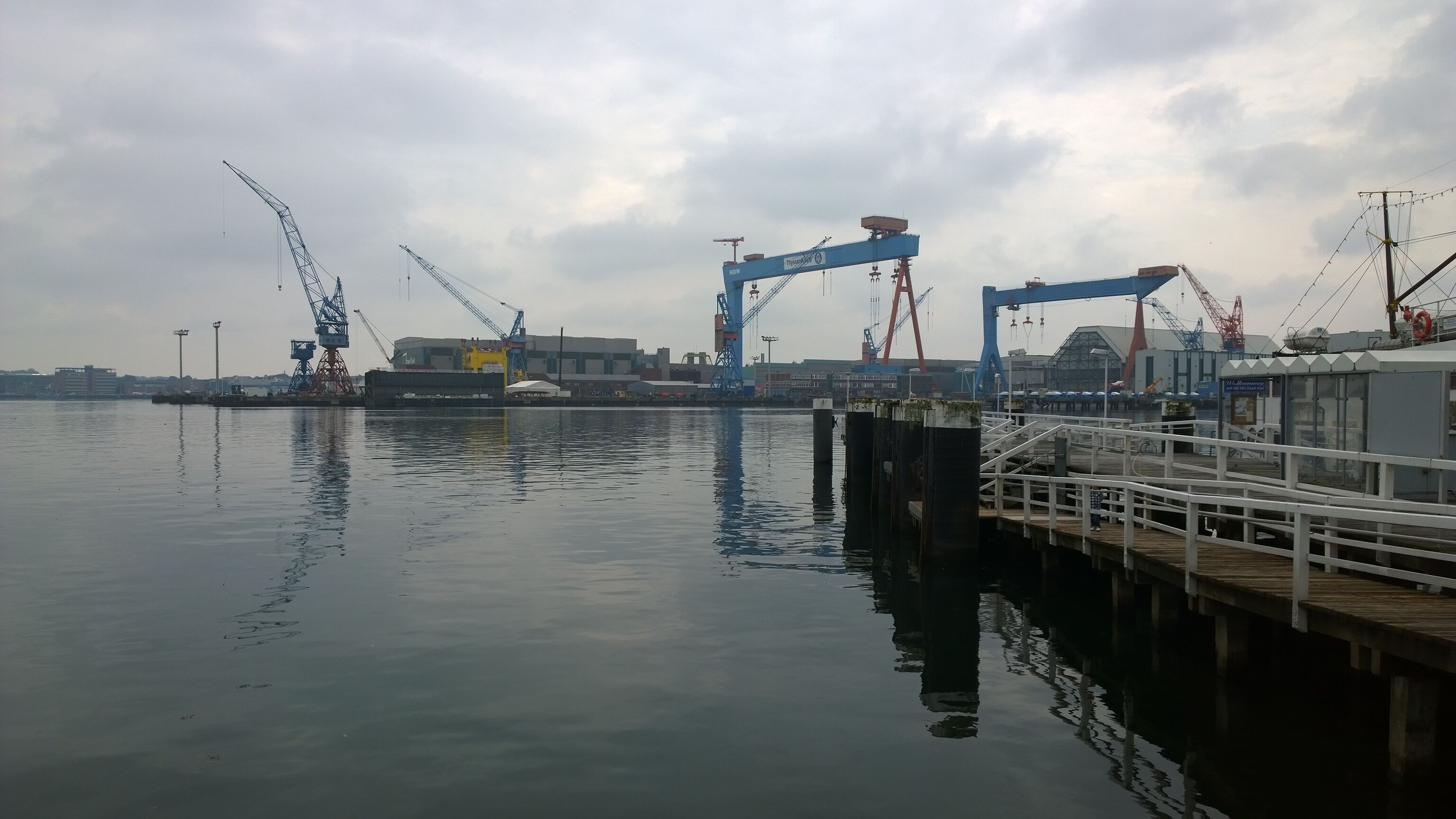 Un moll al costat del mar amb un pontó que s'endinsa en aigües tranquil·les, amb grues industrials i un edifici gran al fons.