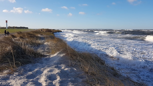 Une plage où les vagues déferlent sur le rivage, un phare en arrière-plan et un sentier herbeux menant à l’eau.
