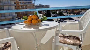 Un balcone con un tavolo apparecchiato con arance e una ciotola di frutta, con vista su una zona costiera, edifici e il mare.