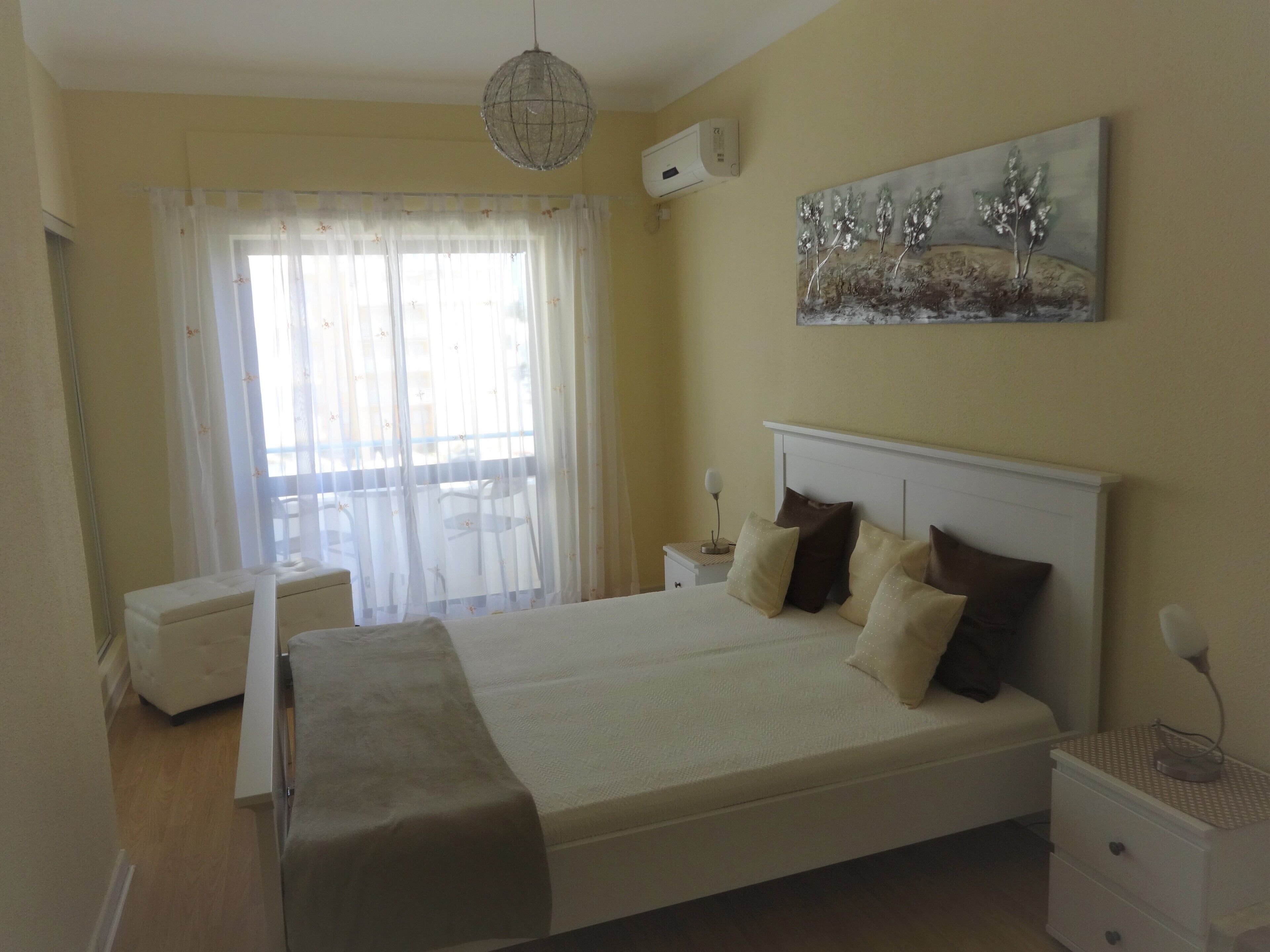 Quarto com cama, mesas de cabeceira, um aparador, janela com cortinas translúcidas e ar condicionado fixado na parede.