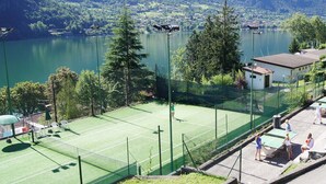 Een tennisbaan met een net, omgeven door een hek, met uitzicht op een meer en bergen.