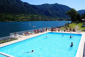 Une piscine où des gens nagent et prennent des bains de soleil, le tout entouré de montagnes et d’un lac.