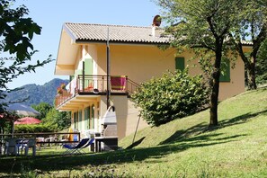 Un bâtiment à deux étages doté d’un balcon, d’un banc en pierre et, au fond, d’une colline verdoyante.