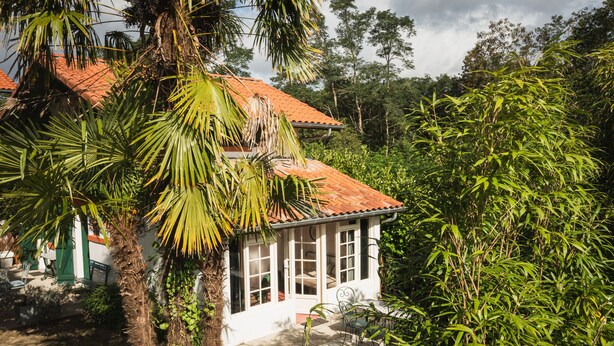 A small white house with a tiled roof, surrounded by lush greenery and trees. - Restored holiday house with all comforts near the sea (Magescq)