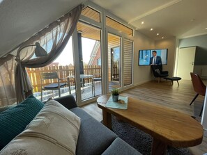 Ein modernes Wohnzimmer mit einer Couch, einem Holzesstisch, einem Fernseher und einem Balkon mit Gartenmöbeln.