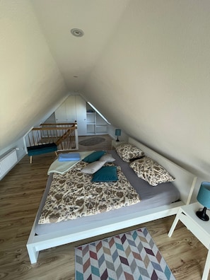 Ein Schlafzimmer mit schräg verlaufender Decke, einem Bett mit gemusterten Betttextilien, einem blauen Sessel und einem geometrischen Teppich.