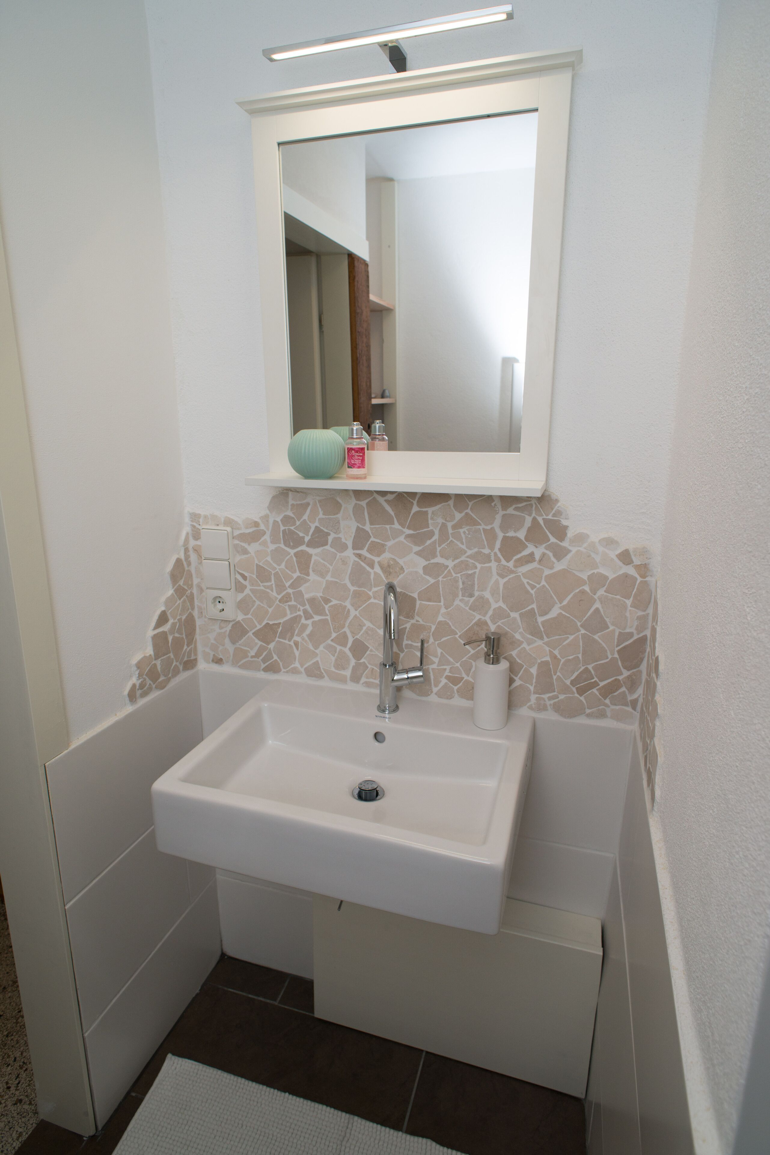 Casa de banho com uma pia branca, espelho e painel de azulejos mosaicados.