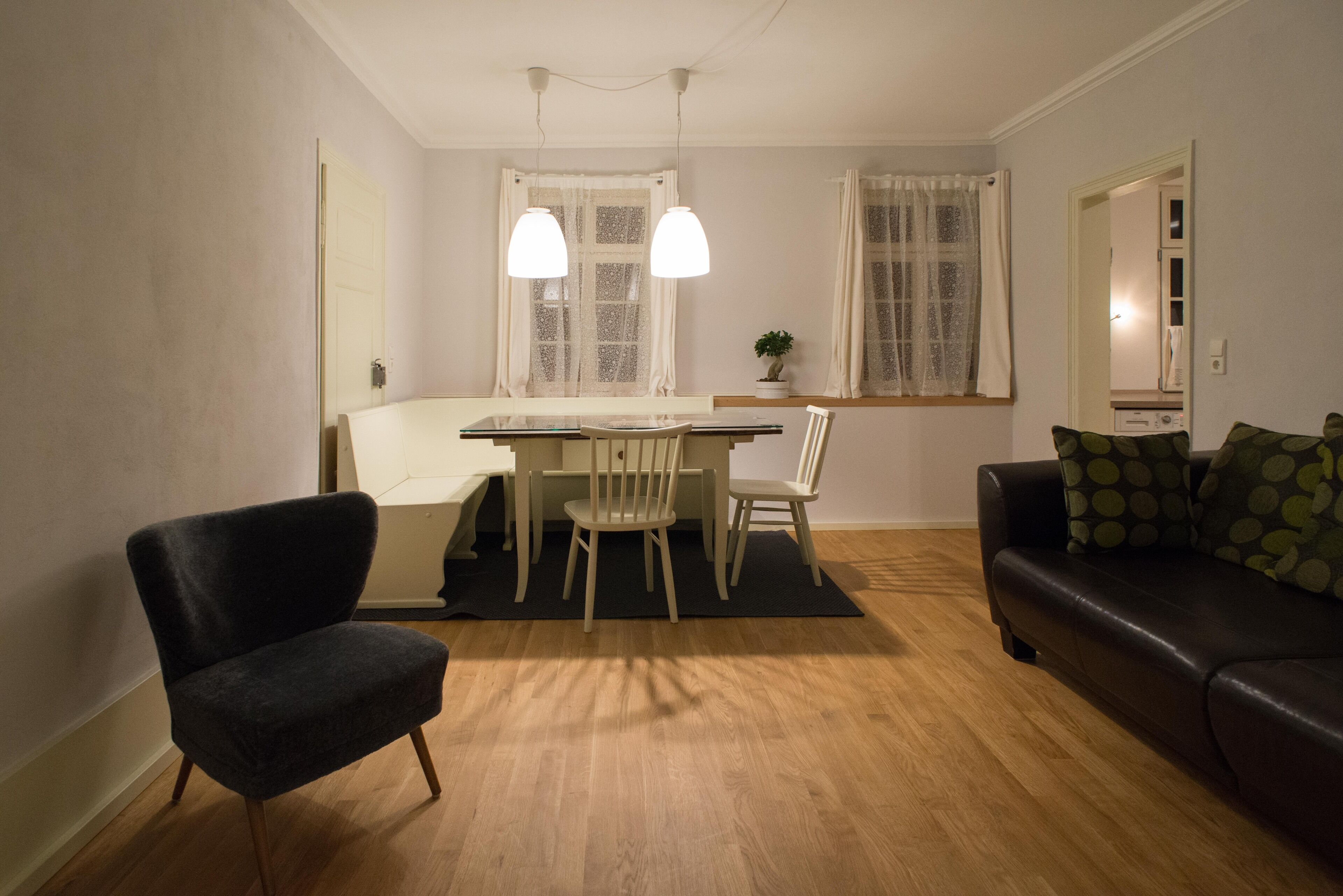 Un saló modern amb un butac armchair fosc, una taula de dinar de fusta amb cadira, un sofà negre amb coixins estampats i una planta en test amb safata a la finestra.