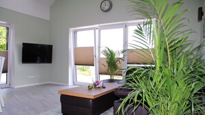 Una sala de estar con televisor de pantalla plana, una mesa de centro de madera, un sofá oscuro, una planta grande en maceta y vistas al exterior a través de una puerta de vidrio.