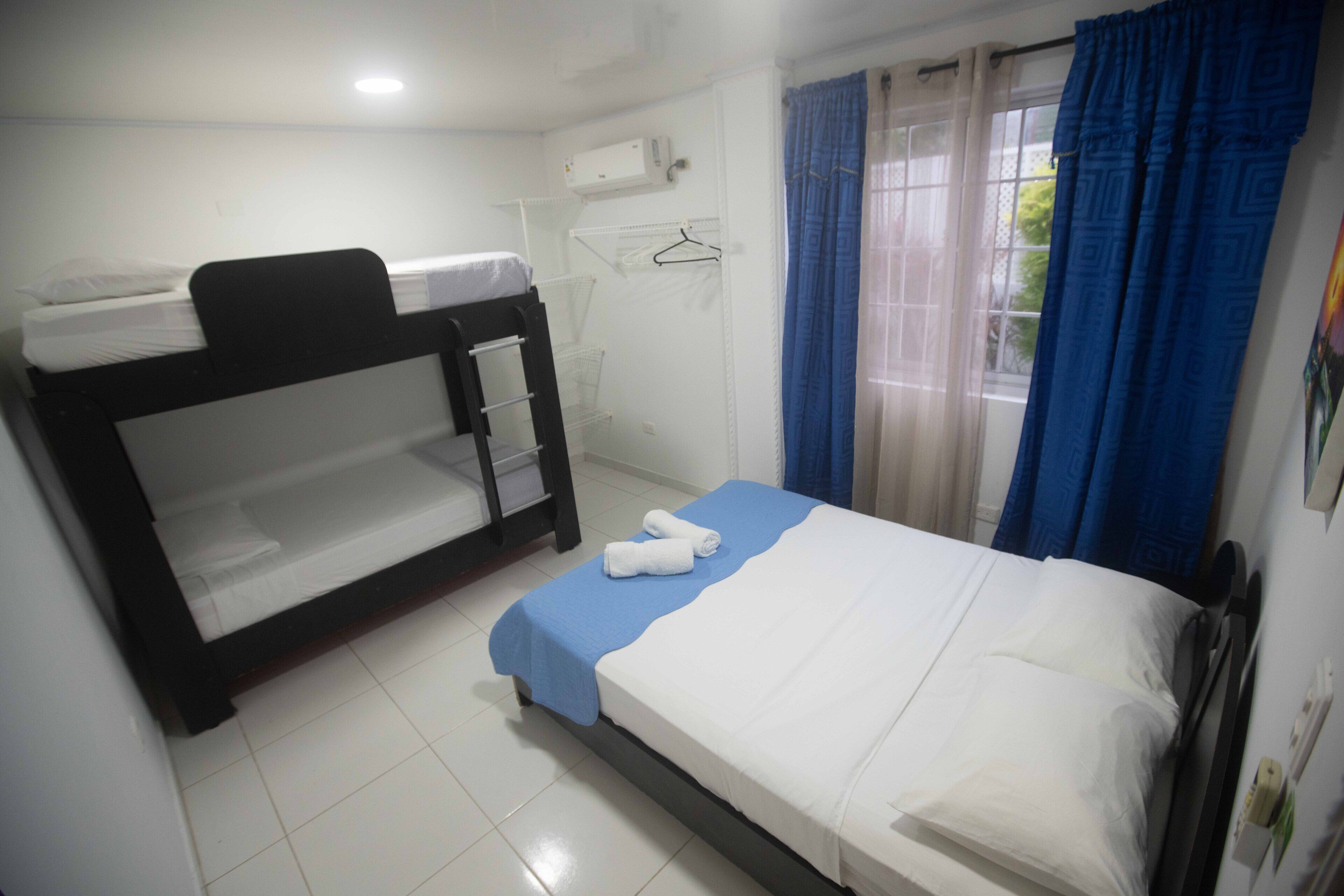 A hotel room with a bunk bed, a single bed, a blue towel on the single bed, a white air conditioner, and a window with blue curtains.