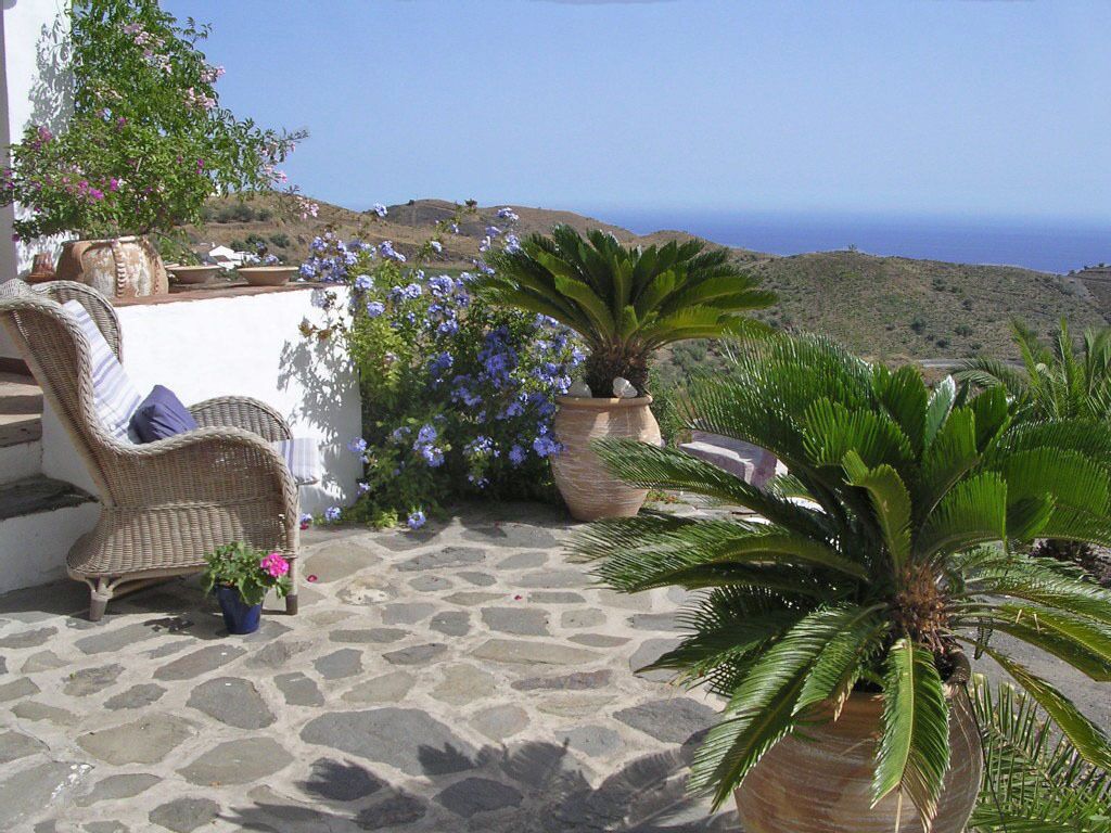 A stone-paved patio with wicker furniture and potted plants, overlooking a hilly landscape and the sea.