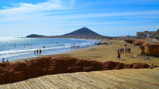 Una playa con un paseo marítimo de madera, gente caminando y una montaña al fondo.