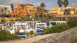 Un hotel frente a la playa con varias zonas de descanso, una pared de piedra y edificios de colores al fondo.