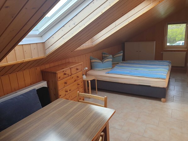A small, cozy room with a sloped ceiling, wooden walls, a bed with striped bedding, a small table, and a chair.