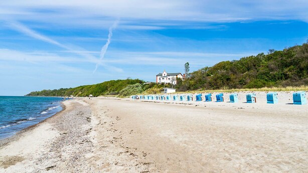 Una playa con una hilera de cabañas azules, una casa grande en una colina y un cielo despejado.