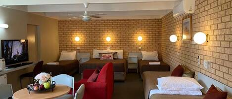 Quarto de hotel com parede de tijolos, duas camas, uma TV, área de refeições com mesa e cadeiras, e um ventilador de teto.