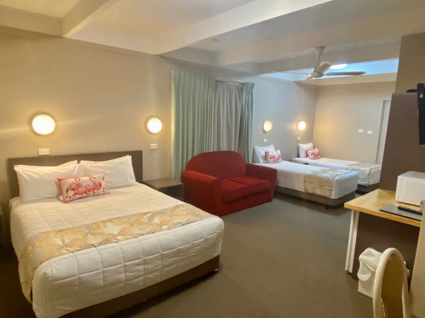 A hotel room with a large bed, a smaller bed, a red armchair, a desk with a computer, and a ceiling fan.