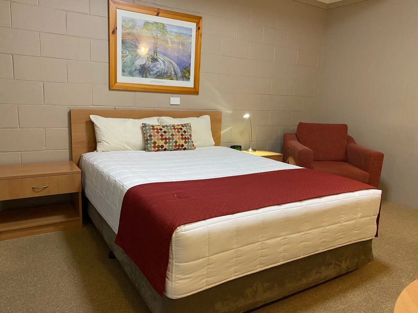 A neatly arranged bedroom with a large bed, a wooden nightstand, a red armchair, and a painting on the wall.