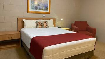 A neatly arranged bedroom with a large bed, a wooden nightstand, a red armchair, and a painting on the wall.