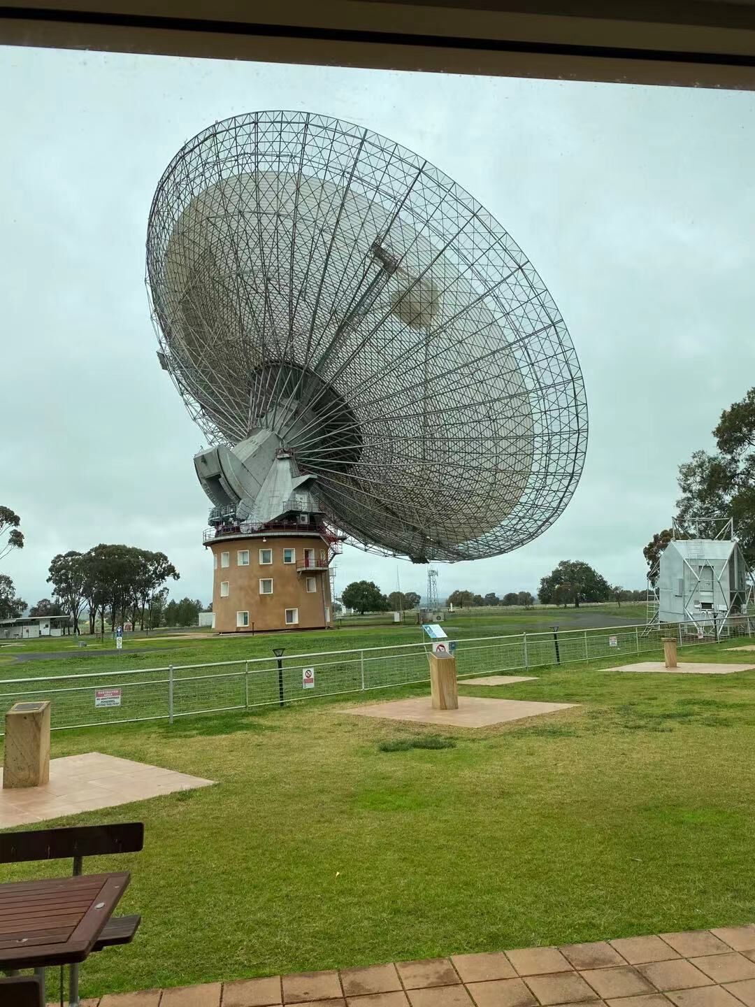 Una gran estructura de antena parabólica con base de edificio, rodeada por un área cercada con césped y árboles.