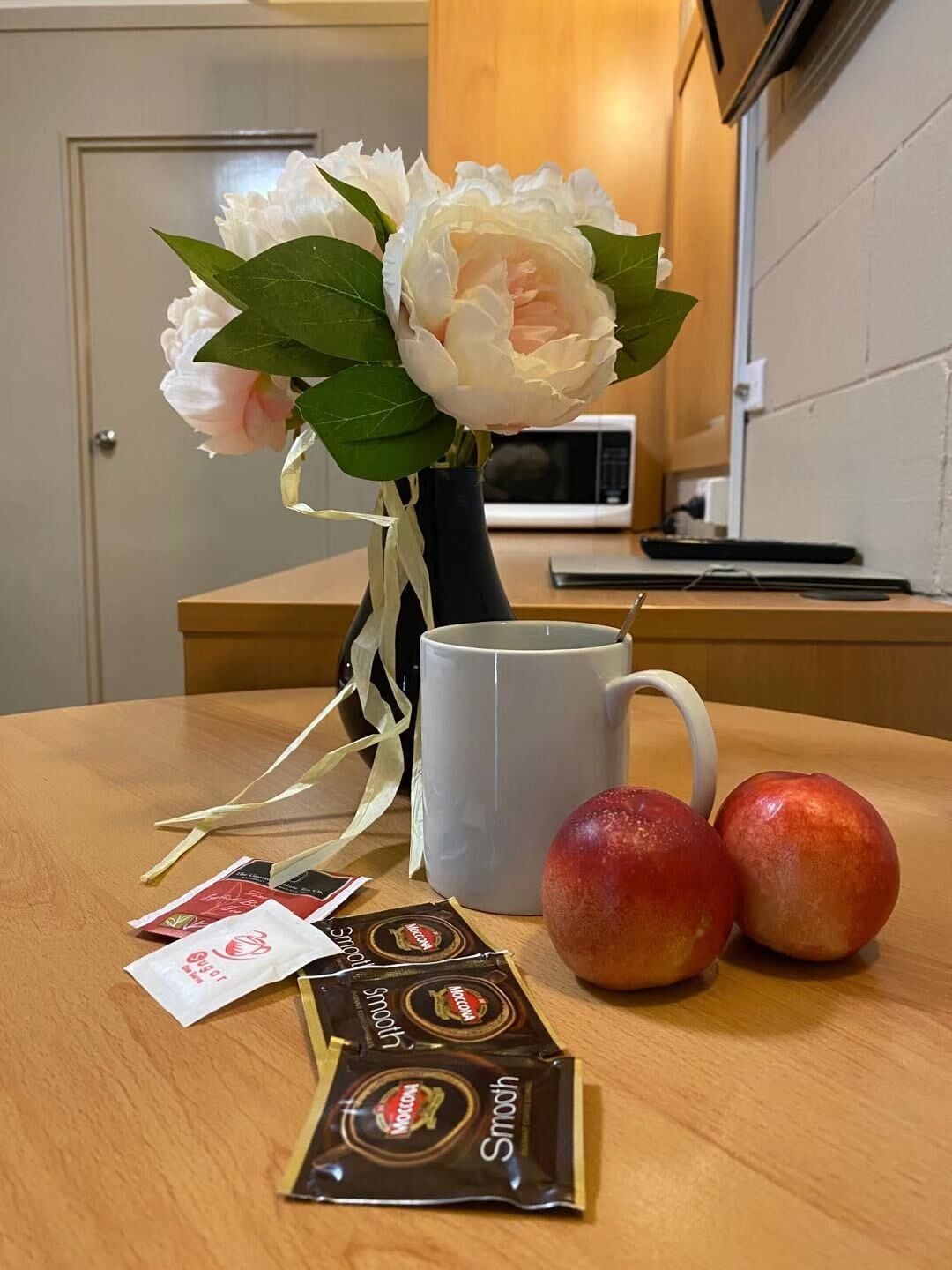 A wooden table with a white mug, two apples, a bouquet of flowers, and chocolate packets.