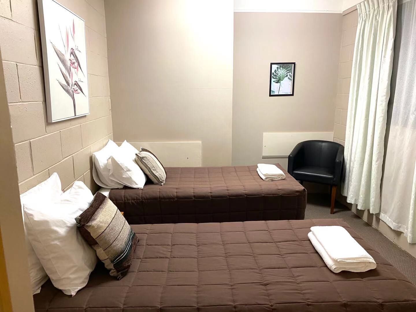 Een kleine, verzorgde kamer met twee bedden, een stoel en een enkel fotokader aan de muur.