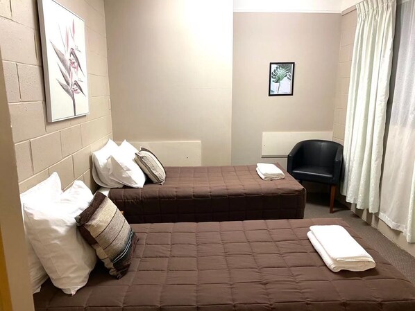 A small, neatly arranged room with two beds, a chair, and a single framed picture on the wall.