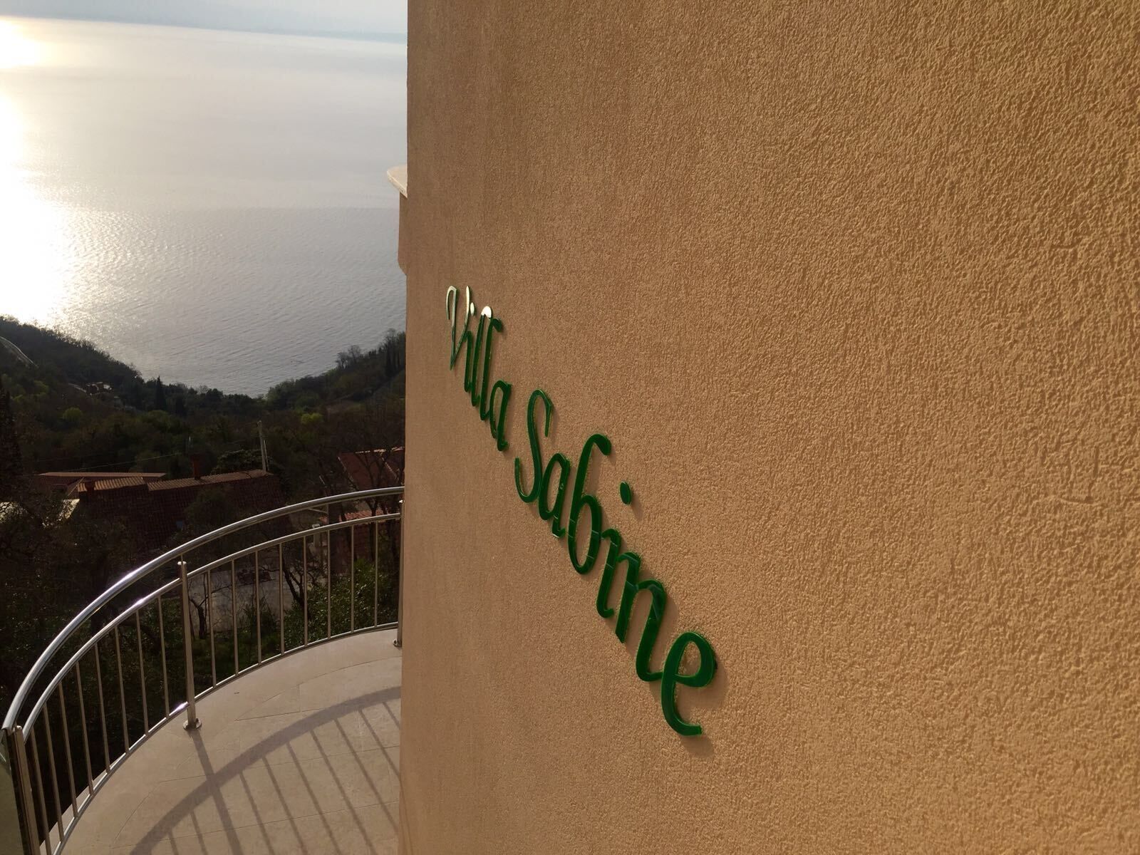 En skylt med texten ”Villa Sabine” sitter på en beige vägg, och i bakgrunden syns havet och kullarna.