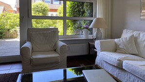 Een kamer met een witte fauteuil, een witte bank en een glazen salontafel. Er is een raam met zonwering, een radiator en een lamp op een bijzettafeltje.