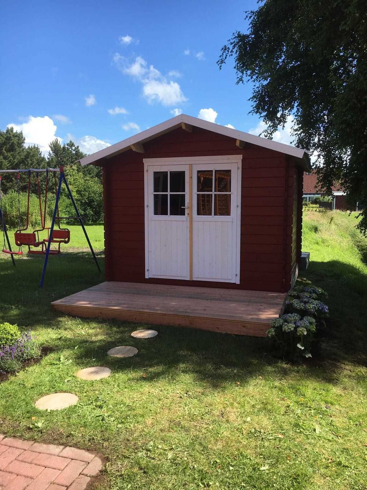Un cobertizo de madera pequeño y rojo con puerta y ventanas blancas, una terraza de madera y un columpio al fondo.