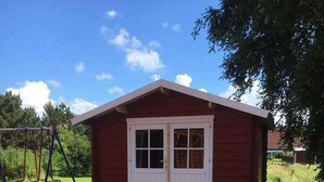 A small red wooden shed with a white door and windows, a wooden deck, and a swing set in the background.