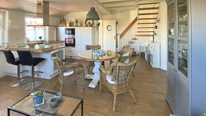 Een ruime woonkamer met een eettafel omringd door rieten stoelen, een glazen salontafel en een keuken met witte kasten en een inbouwoven.