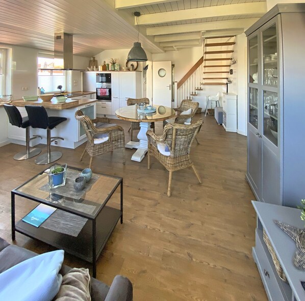 A spacious living area with a dining table surrounded by wicker chairs, a glass coffee table, and a kitchen area with white cabinets and a built-in oven.