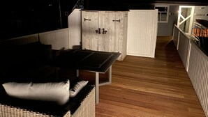 Een overdekt buitengebied met een tafel en stoelen, een houten terras en een wit gebouw met een raam op de achtergrond.