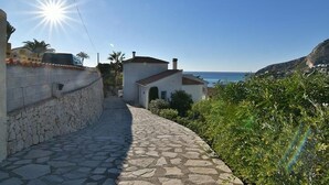 A narrow stone pathway leading to a white building with a red roof, overlooking a coastal area with a clear sky and the sea in the background. - Peaceful Villa with heavenly sea view and panoramic view (Calp)