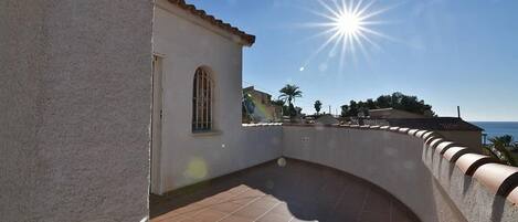 A courtyard with a tiled floor, a small building with a window, and a clear view of the sea under a bright sun.