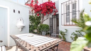 A patio with a metal table and chairs, a white wall with a decorative light, and a red flowering plant.
