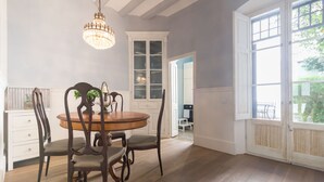 A dining area with a round table, chairs, and a chandelier. There is a window with a view of greenery outside.