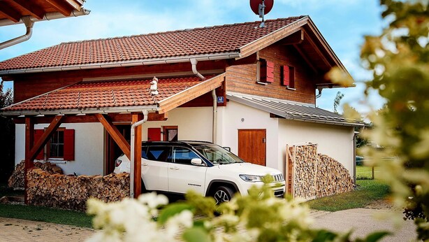 Una casa con techo de tejas, un SUV estacionado y una pila de leña afuera.