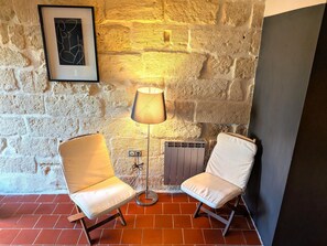 A corner room with a stone wall, a framed abstract artwork, a floor lamp, two white cushioned chairs, and a radiator.