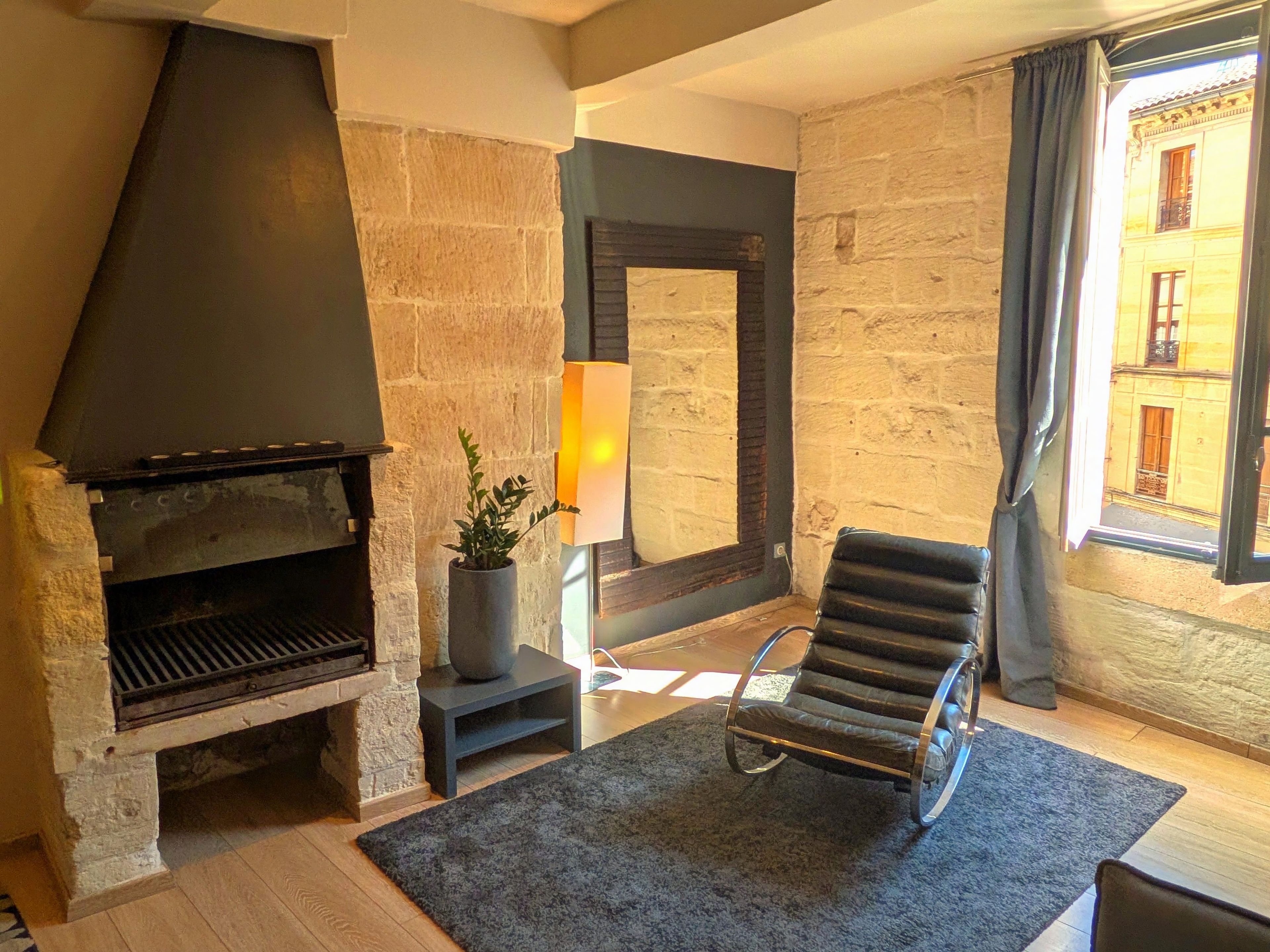 A living room with a stone fireplace, a modern chair, and a view of a building through a window.