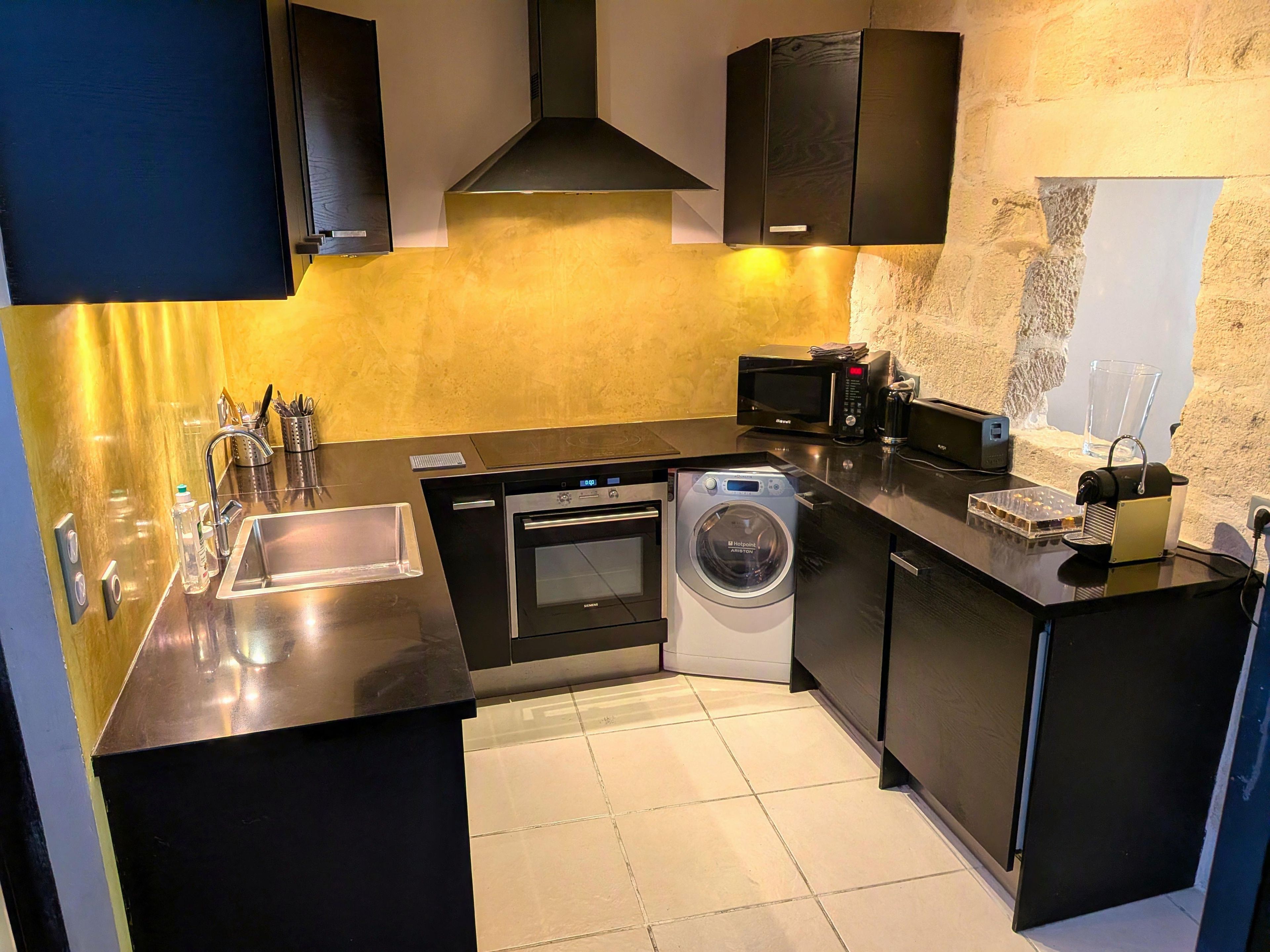 A compact kitchen with a dishwasher, oven, and washing machine, a sink with utensils, and a microwave on the counter.