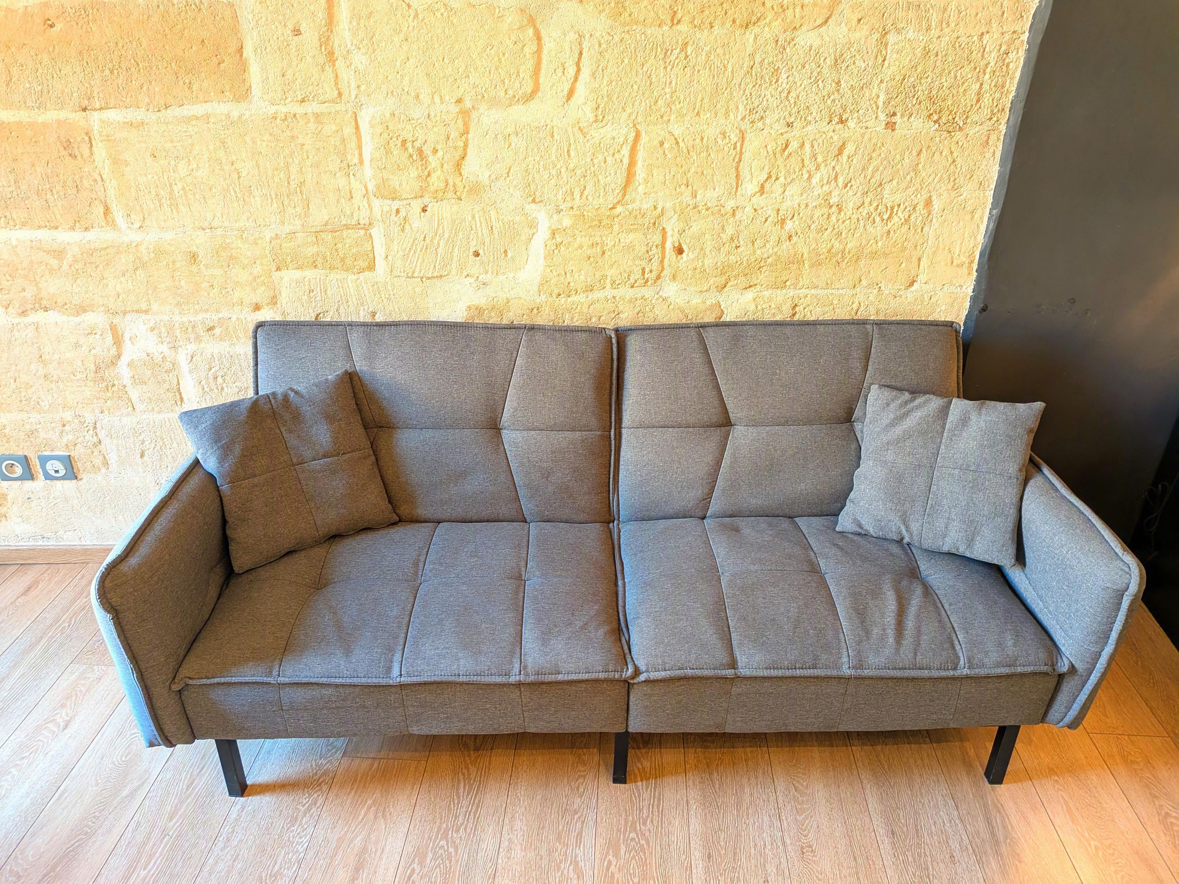 A grey sofa with armrests and cushions against a brick wall.